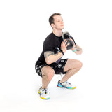 Wearing a black T-shirt and Bells of Steel Gym Shorts, a person in colorful sneakers and with tattoos visible on their arms holds a kettlebell close to their chest while squatting against a plain white background.