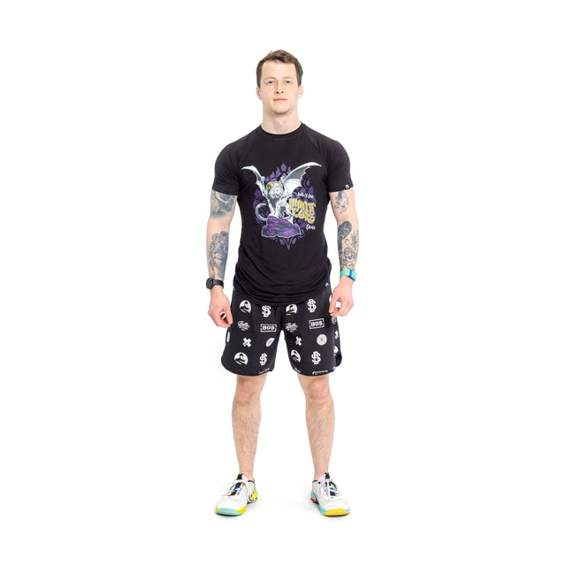 A person in a black T-shirt adorned with a colorful graphic, Bells of Steel gym shorts displaying diverse white symbols, and white sneakers with yellow accents stands against a plain white backdrop.