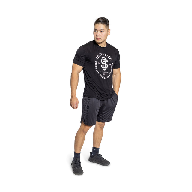 A man stands against a white background wearing Bells of Steel gym shorts and a black T-shirt with a circular logo. He gazes to the side, hands relaxed by his sides, dressed in black socks and sneakers.