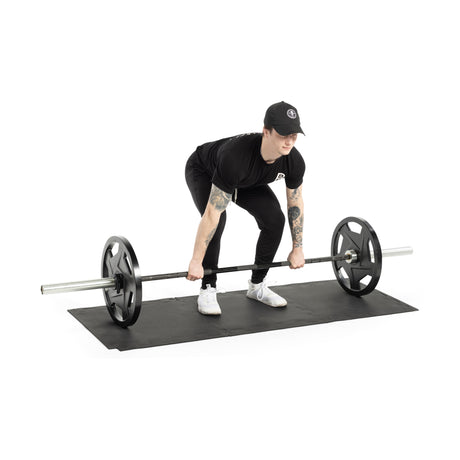 A person in black athletic clothing and a cap prepares to lift a barbell on Bells of Steel 24" x 24" Puzzle Mats. With tattoos on their arms, they're bent over gripping the bar for stability, ensuring the gym floor is protected.