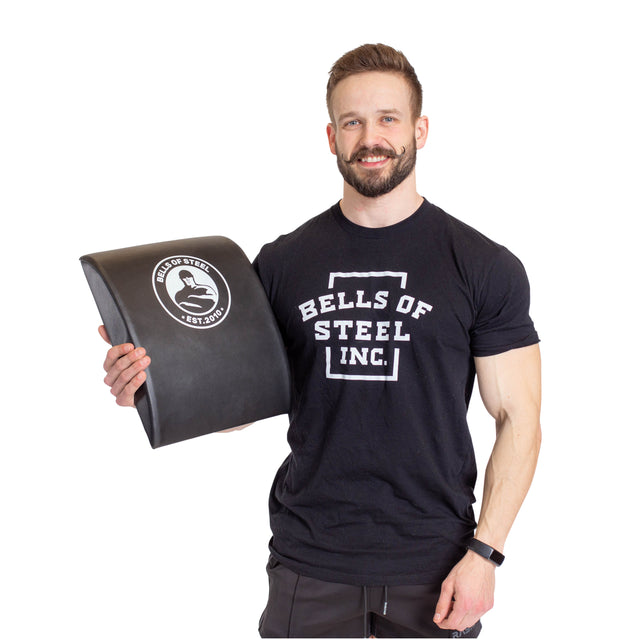 A bearded man smiles, holding a large Sit Up Mat with a logo. He wears a black Bells of Steel T-shirt and gray shorts against a white background. The mat features lumbar support for core workouts.