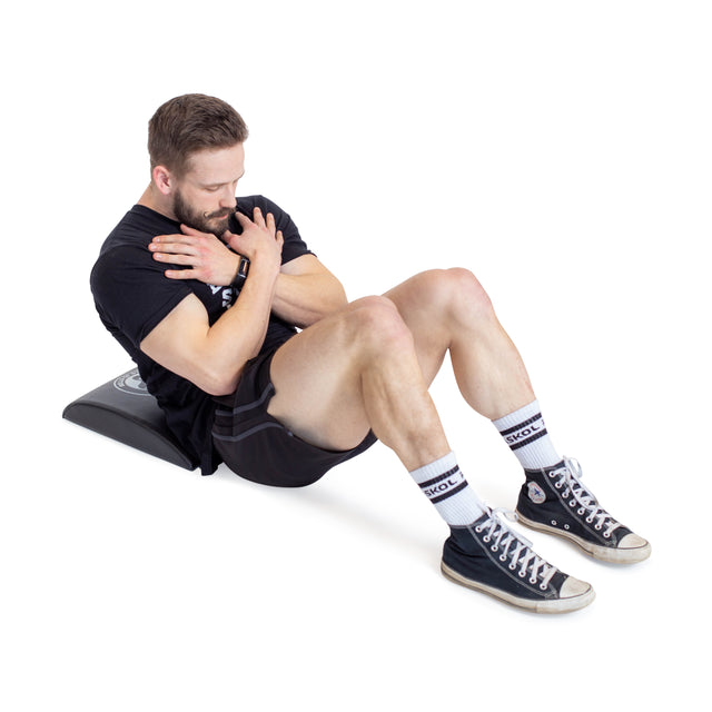 A bearded man in a black T-shirt, shorts, white socks, and black high-tops focuses on his core workout. He performs sit-ups on the Bells of Steel Sit Up Mat with lumbar support, arms crossed and eyes closed against a plain white backdrop.