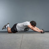 A man with curly hair kneels on a gym floor, wearing a gray t-shirt and black shorts, using the Bells of Steel ABsyidyan Stainless Steel Ab Roller with an extra-wide dual wheel to extend his body forward.