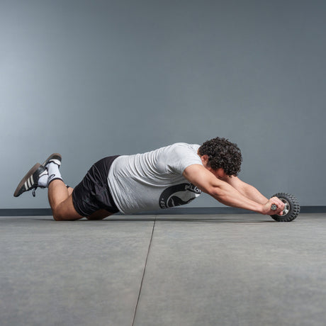 A man with curly hair kneels on a gym floor, wearing a gray t-shirt and black shorts, using the Bells of Steel ABsyidyan Stainless Steel Ab Roller with an extra-wide dual wheel to extend his body forward.