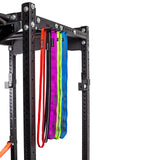 Four resistance bands in red, black, purple, and blue hang from a Bells of Steel Accessory Holder (Wall or Hydra Mounted) attached to a black metal power rack, providing efficient gym storage against a white background.