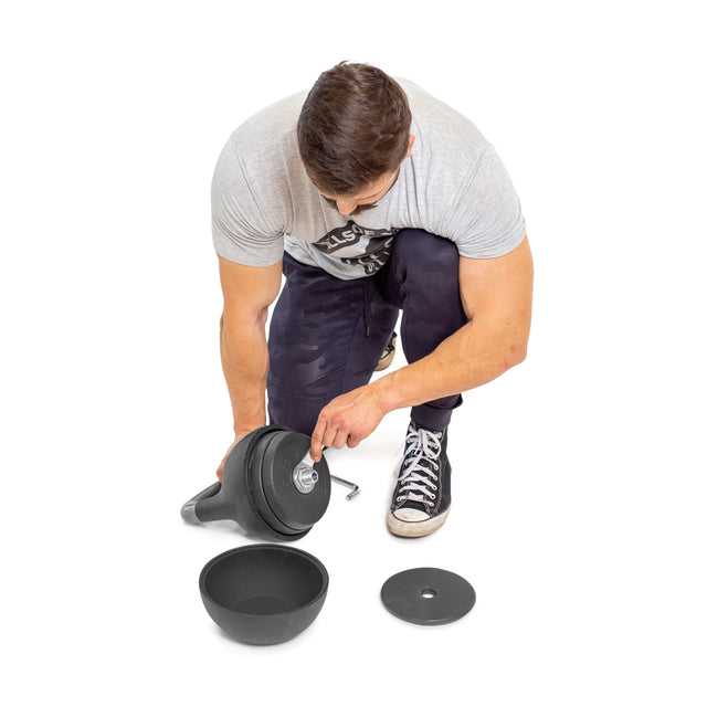 A person is kneeling, adjusting a Bells of Steel Adjustable Competition Kettlebell. Some weight increments and a disc are on the floor. They wear a gray shirt, black pants, and black and white sneakers.