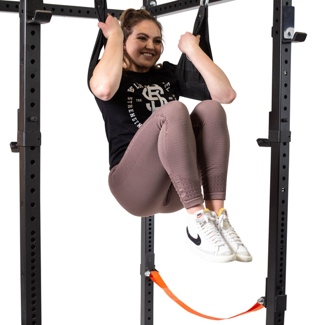 A woman is working out on a pull-up bar, holding her legs up with the support of Bells of Steel Hanging Ab Straps. She smiles confidently in a black T-shirt, pink leggings, and white sneakers featuring the black Nike logo. The heavy-duty carabiner ensures her Hanging Ab Straps remain securely fastened.