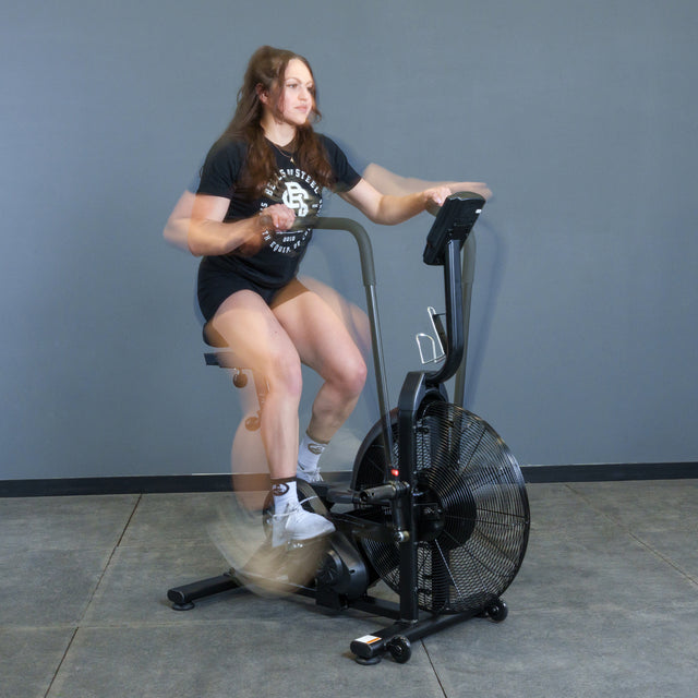 A woman rides the Bells of Steel Residential Air Bike with Wind Guard indoors. Her quick movements blur against a minimalist home gym backdrop with a gray wall and concrete floor.