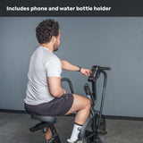 A man in athletic wear uses the Bells of Steel Residential Air Bike with Wind Guard, adjusting his phone in the built-in holder. Text at top: "Includes phone and water bottle holder"—ideal for your home gym setup.
