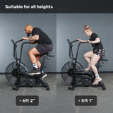 Two people—one 6'2" and one 5'1"—ride the Bells of Steel Residential Air Bike with Wind Guard together, proving it suits users of any height. Text above reads, "Suitable for all heights." Perfect addition to any home gym.