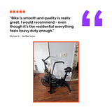 A Bells of Steel Residential Air Bike with Wind Guard sits on a wooden floor in a modern home gym, alongside a customer review praising the fan bike’s smoothness, quality, and durability for residential use.