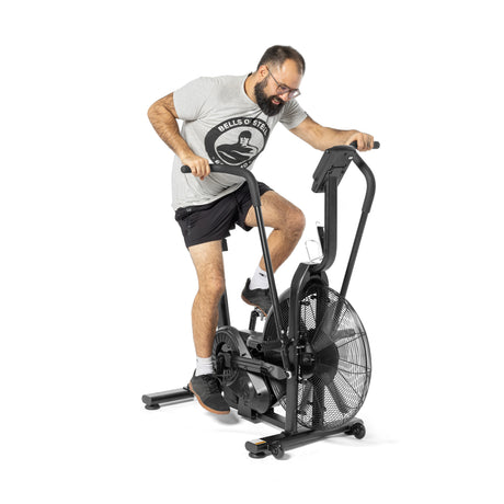 A man exercises on a Bells of Steel Residential Air Bike with Wind Guard in his home gym, wearing a gray T-shirt, black shorts, and black sneakers. The bike includes a large fan wheel and sturdy handlebars.