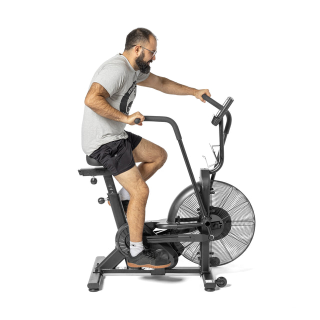 A bearded man with glasses works out vigorously on a Bells of Steel Residential Air Bike with Wind Guard, dressed in a gray T-shirt, black shorts, and black shoes. The bike's large front fan spins rapidly against the basic white backdrop, showcasing his commitment to his home fitness regimen.