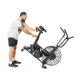 A man in a gray T-shirt and black shorts adjusts the seat of a Bells of Steel Residential Air Bike with Wind Guard. Wearing glasses and sporting a beard, he stands on a white background, embodying the essence of a modern home gym workout.