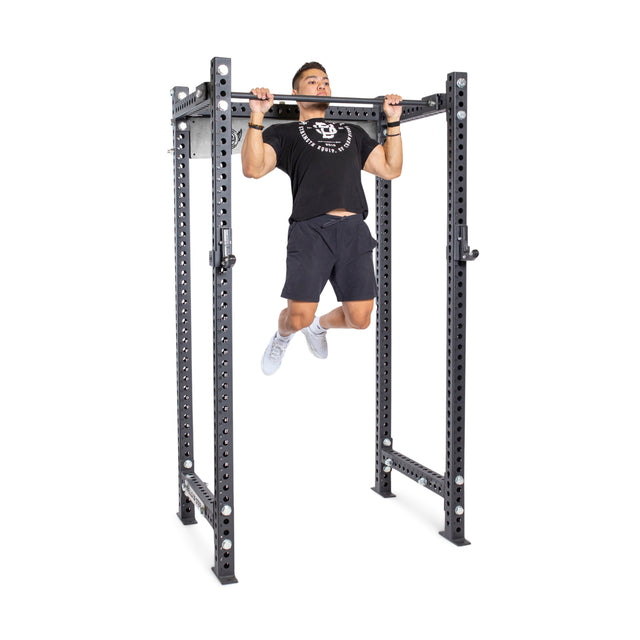 A man performs a pull-up on the Bells of Steel Adjustable Pull-up Bar Rack Attachment. He wears a black t-shirt, black shorts, and white sneakers. The metal frame has multiple holes along the sides for easy adjustments.