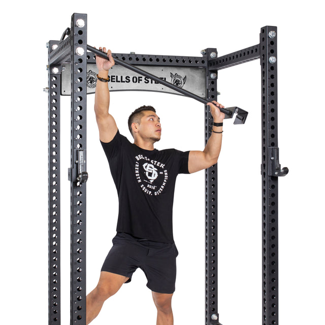 A person in a black t-shirt and shorts assembles an Adjustable Pull-up Bar Rack Attachment by Bells of Steel, holding a bolt in their left hand and a tool in their right to secure a component, while the pull-up bars lie nearby.