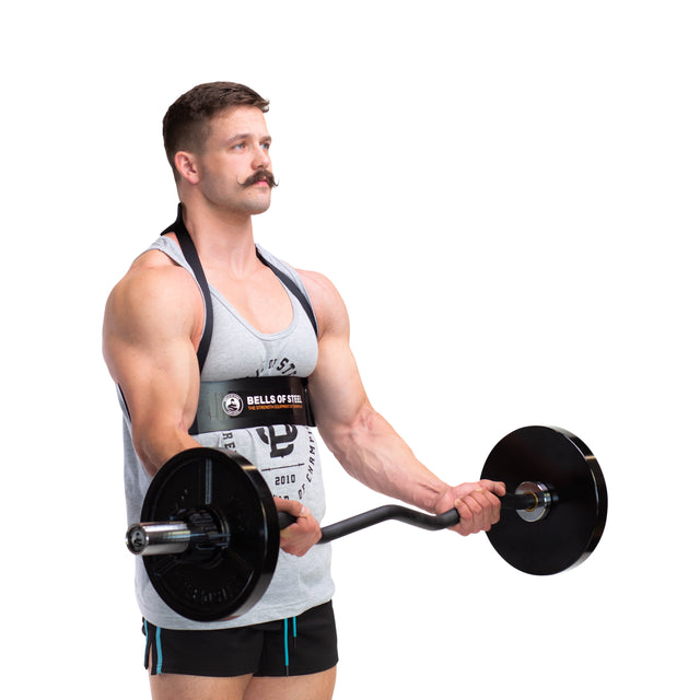 A man with a mustache is lifting a barbell with weights, maintaining proper form. He wears a gray tank top and black shorts, standing against a white background. A Bells of Steel Arm Blaster, recognized for its durable aluminum build, is strapped around his upper body to support his arms.