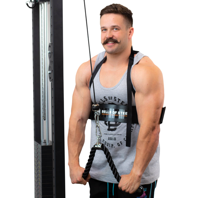 A man with a mustache demonstrates an upper body exercise, emphasizing proper form using the Arm Blaster from Bells of Steel. He is dressed in a gray tank top and black shorts, firmly holding a rope attachment against a plain white backdrop.