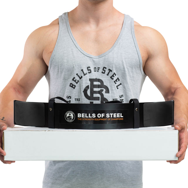 A person wearing a gray tank top with "Bells of Steel" in black lettering is holding a white box containing an Arm Blaster, which features the Bells of Steel branding. The accompanying belt promotes itself as the "Bells of Steel: The Strength Equipment of Champions," designed to help maintain proper form.