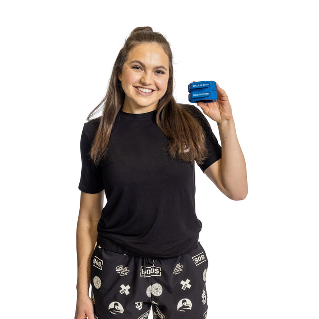 A smiling woman with long brown hair wearing a black t-shirt and patterned black pants holds up a Bells of Steel Magnetic Clamp Collar against a white background.