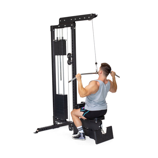 A person in a gray tank top and black shorts uses the Bells of Steel Lat Pulldown Bar at a cable machine, performing lat pulldowns. They pull the bar toward their chest, engaging their muscles as the weights stack on the machine.