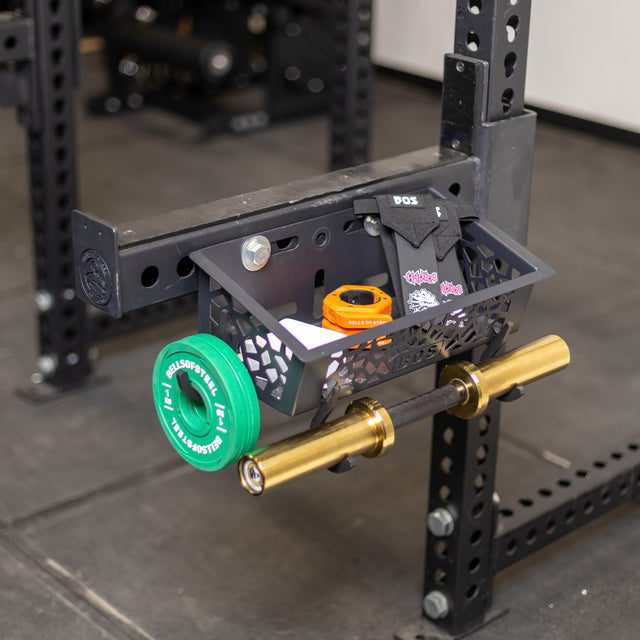 A Bells of Steel Rack Attached Storage Basket holds wrist wraps, a notepad, and a tape measure. A mini barbell with green weight plates and orange athletic tape are conveniently stored on a horizontal rack attachment nearby.