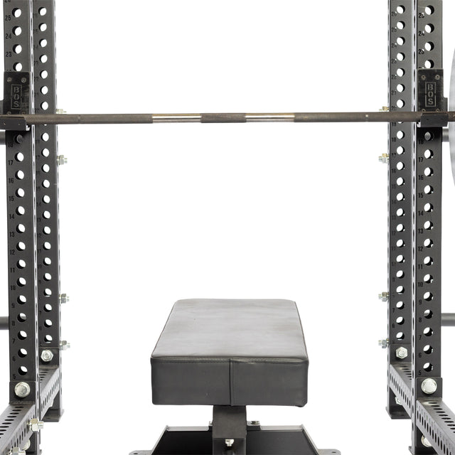 A weight bench set beneath a barbell, securely rested on Standard J-Cups by Bells of Steel within a power rack. The equipment is black and sturdy with UHMW protection, making it ideal for strength training in a gym or home setting.