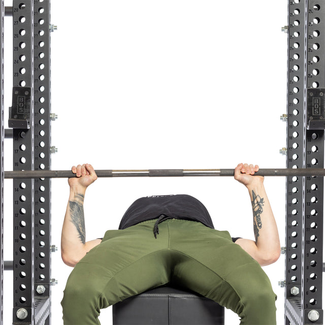 Wearing olive green pants, a person is performing a bench press on a bench, lifting a barbell within a white background. Their tattooed arms flex with effort inside a power rack equipped with Bells of Steel Standard J-Cups featuring UHMW protection.