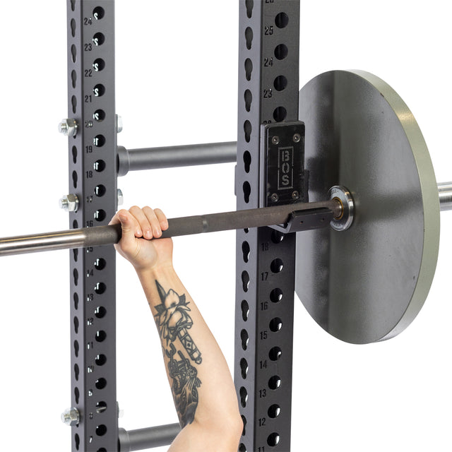 A person with a tattooed arm grips a barbell on a power rack lined with UHMW protection. The black Bells of Steel squat rack features Standard J-Cups and numbered holes for perfect height adjustment.