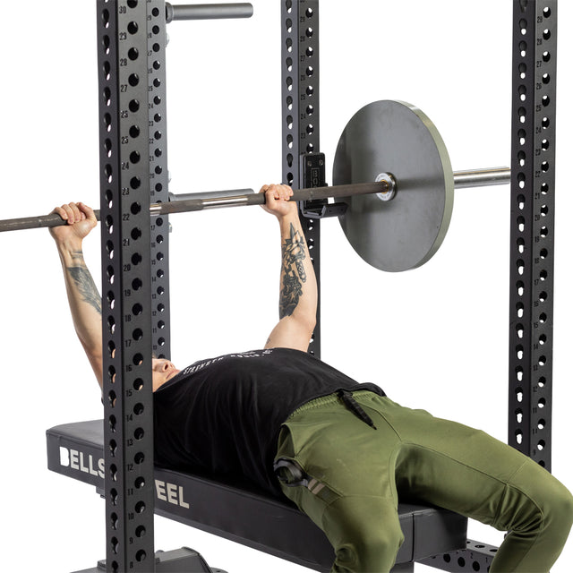 A person in a black shirt and green pants is performing a bench press with a barbell securely resting on the Bells of Steel Standard J-Cups, which are part of an impressive power rack setup. Their tattooed arms flex as they lift with precision and strength.