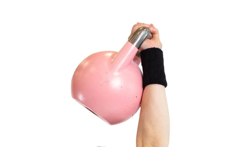 A person, equipped with Bells of Steel's Mighty Wrist Wraps, is lifting a pink kettlebell overhead against a plain white background.