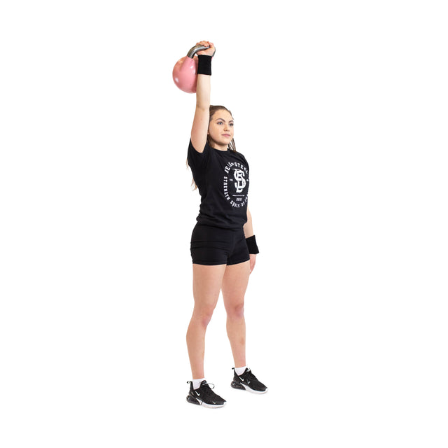 Dressed in a black t-shirt and shorts, a person balances on one leg while holding a pink kettlebell overhead. With sneakers and BellGuard Kettlebell Wrist Guards by Bells of Steel, they stand poised like seasoned lifters against a plain white background.