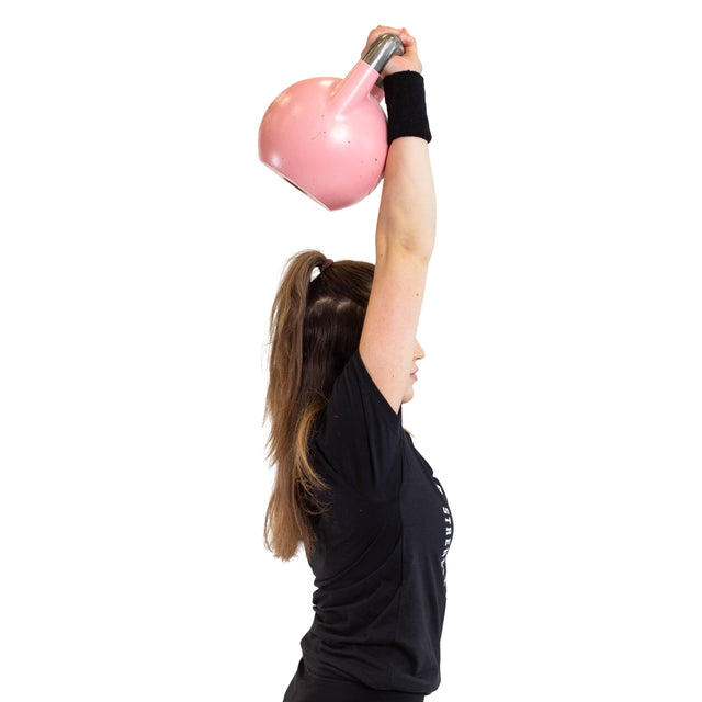 A kettlebell lifter in a black shirt hoists a pink kettlebell overhead with one arm, showcasing strength and balance. With long brown hair and focus against the plain white background, they wear a wristband for support and BellGuard Kettlebell Wrist Guards by Bells of Steel for added protection.