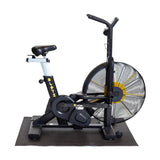 A Bells of Steel Cardio Machine Floor Mat is shown against a white background with a stationary air bike featuring a large fan wheel, dual handlebars, and a padded seat placed on top.