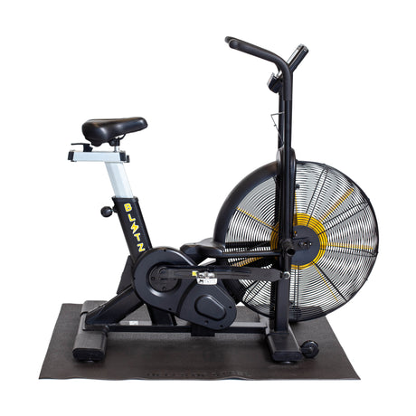 A Bells of Steel Cardio Machine Floor Mat is shown against a white background with a stationary air bike featuring a large fan wheel, dual handlebars, and a padded seat placed on top.