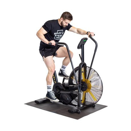 A man in a black T-shirt and shorts works out on an air bike, gripping the handles and pedaling over a Bells of Steel Cardio Machine Floor Mat against a white background.