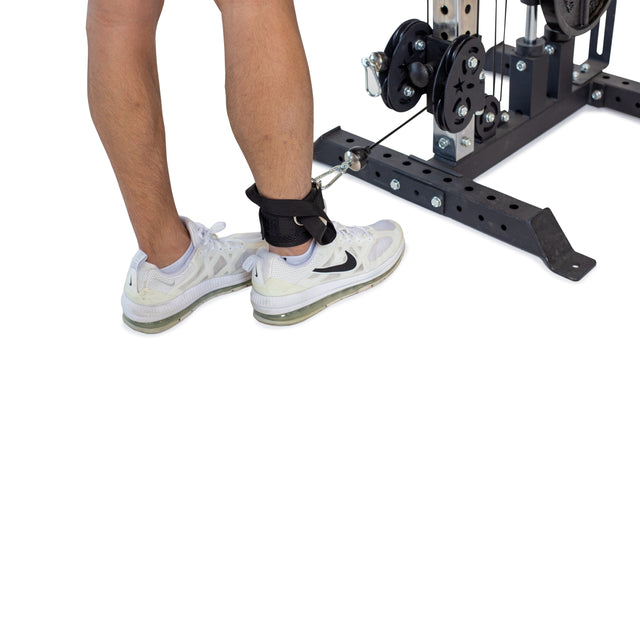 A person with white sneakers and a Bells of Steel Adjustable Ankle Strap Cable Attachment is performing a glute-training leg exercise on a cable machine, highlighting their legs and the gym equipment.