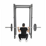 A person in black workout gear and a cap squats with a loaded barbell inside a power rack, using Bells of Steel All-Black Bumper Plate Sets made from recycled rubber, viewed from behind against a white background.