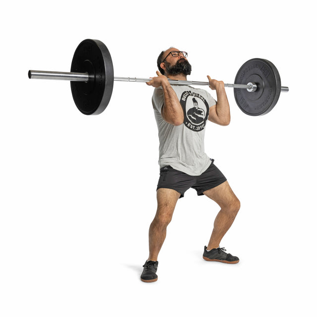 A man in glasses, a grey T-shirt, and black shorts executes a weightlifting move with precise technique. He lifts a barbell at shoulder height in the gym, ensuring safety using All-Black Bumper Plates by Bells of Steel against a plain white background.