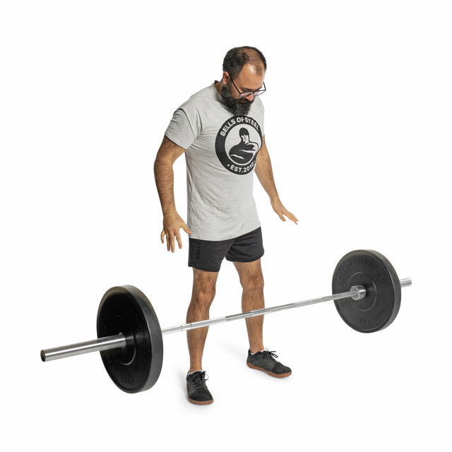 A bearded man with glasses, wearing a light gray T-shirt, black shorts, and sneakers, stands looking down at a Bells of Steel All-Black Bumper Plate Set loaded on a barbell in front of him against a white background.