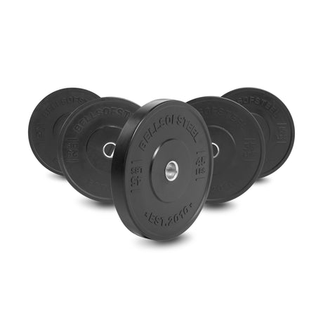 Five Bells of Steel All-Black Bumper Plate Sets are lined up, each with stainless steel insert rings. The front plate displays "45 LBS / 20 KG" and "BELLS OF STEEL," while the other plates show different weights.