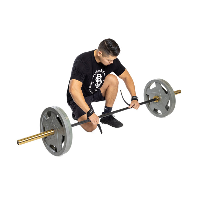 A man kneels beside a barbell, adjusting his Bells of Steel Lifting Straps to enhance his grip strength. He is wearing a black T-shirt with white lettering, black shorts, and wristbands. The barbell features large gray weight plates set against a white background.