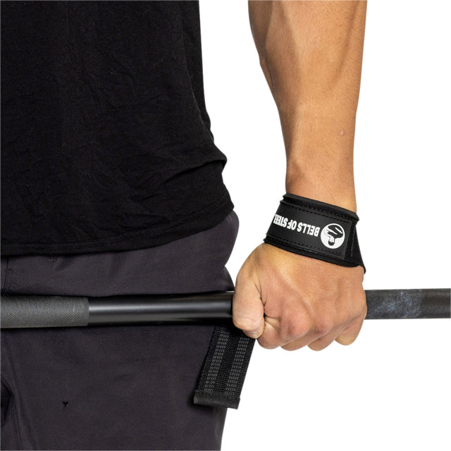 A weightlifter holds a black barbell with their left hand, using a strap from Bells of Steel to improve grip strength. Clad in a black shirt and dark pants, they exude determination and focus.