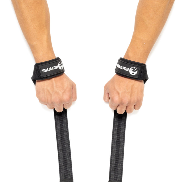 Two hands are displayed with Bells of Steel black lifting straps, each firmly wrapped around a wrist and extending downward. The straps feature the "BELLS OF STEEL" logo in bold white text on a black band. Positioned palm down against a crisp white background, they are perfect for enhancing grip strength for weightlifters.