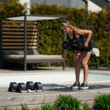 A woman in athletic wear lifts Nuobell NÜOBELL Adjustable Dumbbells with a medium-knurled handle outdoors on a wooden deck, surrounded by machined steel weight plates and greenery.