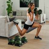 Wearing athletic gear, a woman does a Bulgarian split squat in her living room using Nuobell NÜOBELL Adjustable Dumbbells with medium-knurled handles. Extra Nuobell machined steel weight plates are on the floor; one foot is propped on the couch.