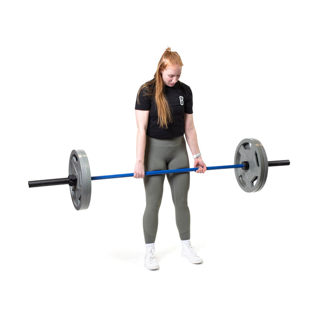 In a pristine, white space, someone in a black shirt and green leggings lifts the Bells of Steel Multi-Purpose Olympic Barbell – The Utility Bar, adorned with gray weights and medium-light knurling. Their long reddish hair is tied back as they wear white sneakers.