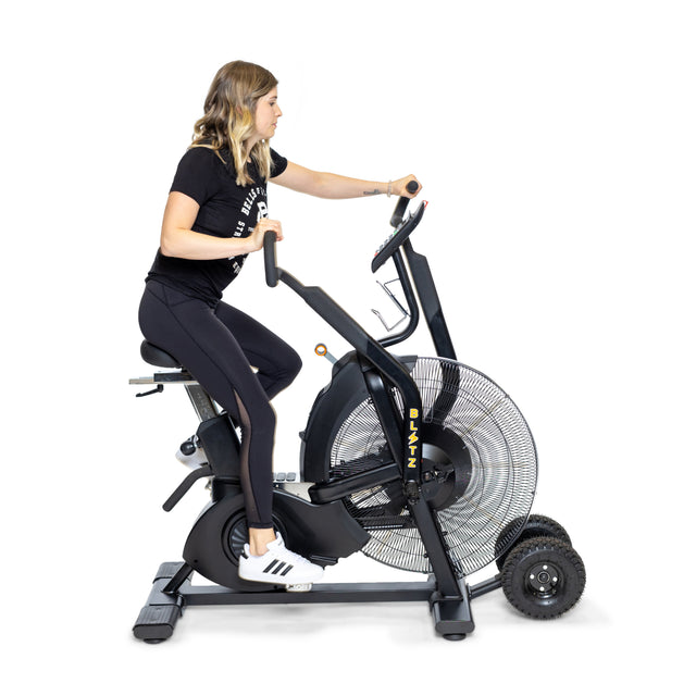 A person exercises on a Bells of Steel Blitz Max Air Bike, wearing black shirt and leggings with white sneakers. The bike is equipped with large fan blades and dual-action handles. Set in a minimalist fitness studio against a plain white background, the scene embodies a commitment to fitness.