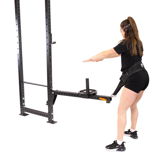 Wearing a black workout outfit, a woman utilizes the Bells of Steel Belt Squat / Lever Arms Rack Attachment, an essential piece for any home gym. She stands with her arms extended forward, harnessed to the versatile component integrated into the robust metal gym structure.