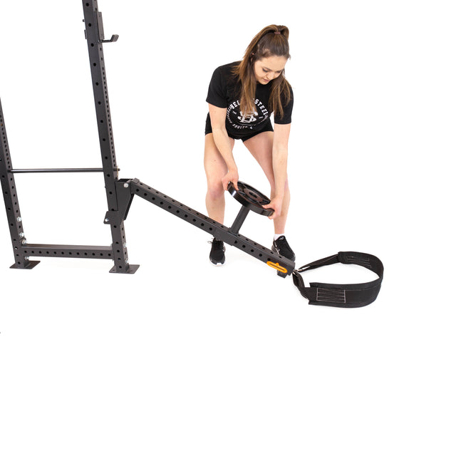 A woman in athletic wear attaches a weight plate to the Bells of Steel Belt Squat / Lever Arms Rack Attachment against a white background. Dressed in a black T-shirt and shorts, her hair is neatly tied up. This equipment is part of a versatile home gym metal rack system.